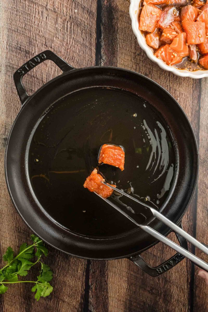 Tongs holding a raw salmon cube over a cast iron skillet with oil, a bowl of marinated salmon and herbs nearby on a wooden surface.