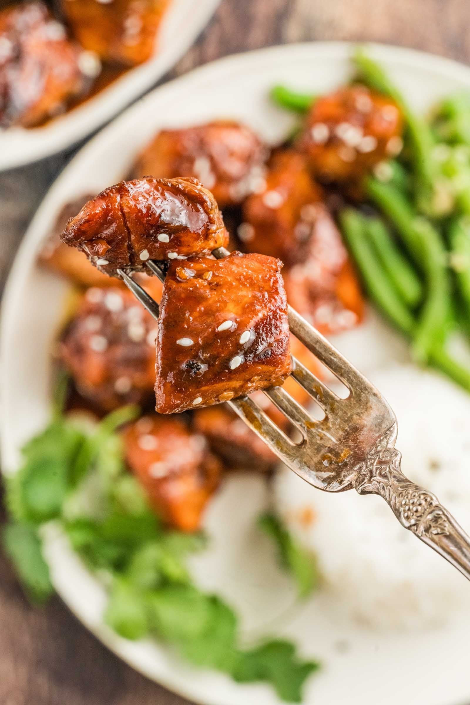 A fork holds a glazed piece of hot honey salmon bites topped with sesame seeds, with rice, green beans, and cilantro visible on a plate in the background.