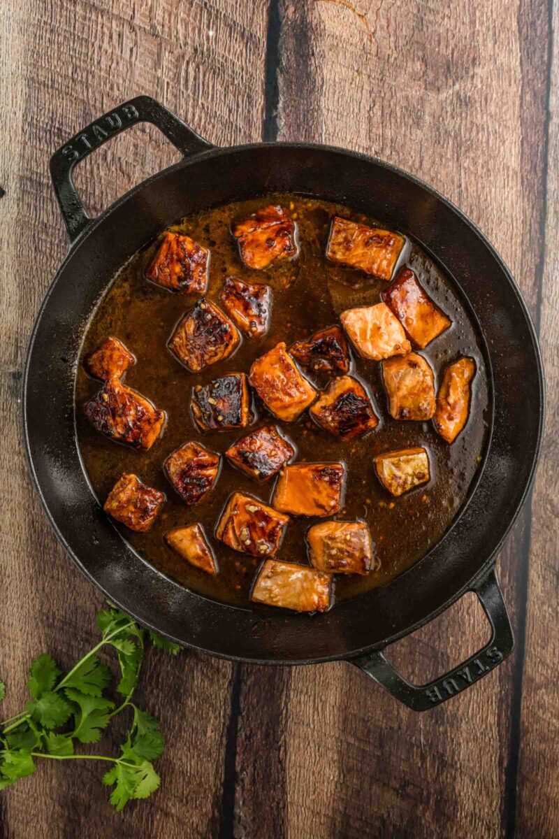 A cast iron skillet with browned salmon bites simmering in a hot honey sauce, placed on a wooden surface with cilantro on the side.