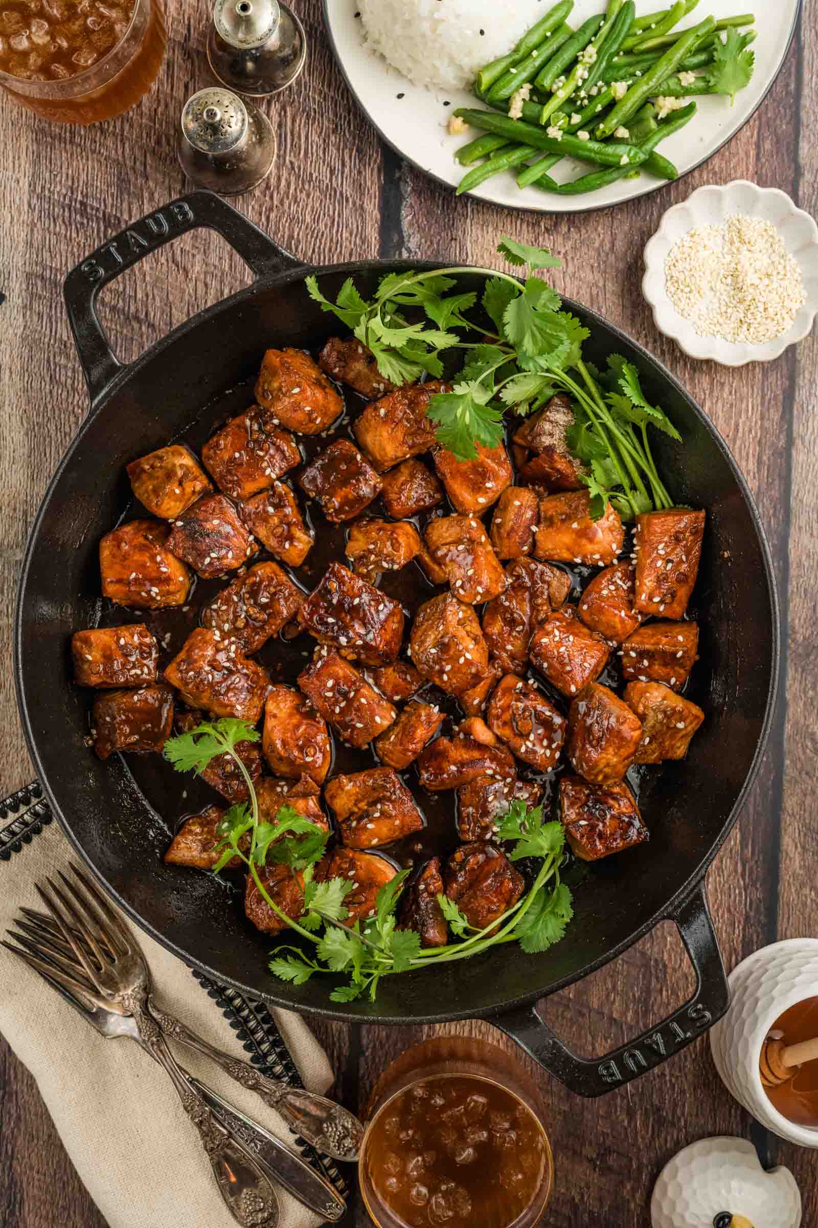 A cast iron pan filled with glazed salmon bites garnished with cilantro and sesame seeds, surrounded by plates of rice, green beans, and drinks on a wooden table.