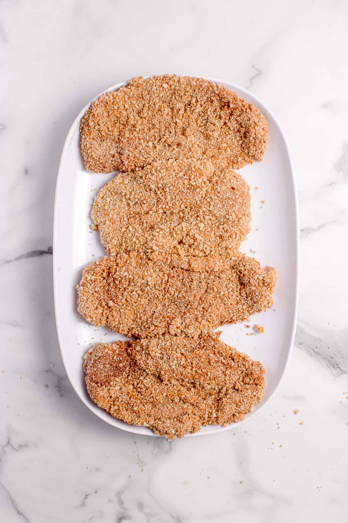 Four breaded cutlets are arranged in a row on a white oval plate set on a marble surface.