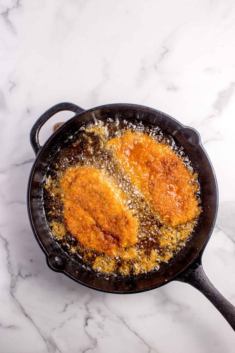 Two breaded cutlets are being deep-fried in oil in a black cast iron skillet on a marble countertop.
