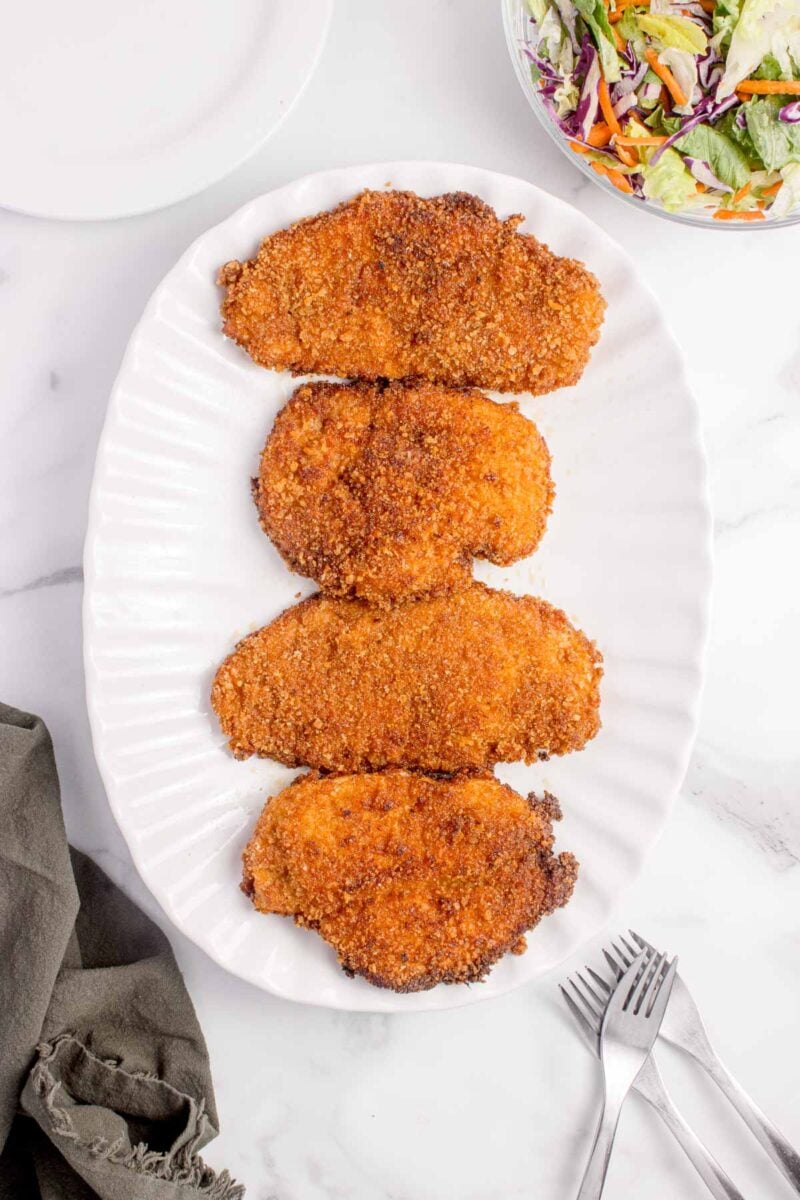 Four breaded and fried chicken cutlets are arranged in a row on a white oval platter, with a bowl of mixed salad and utensils nearby.