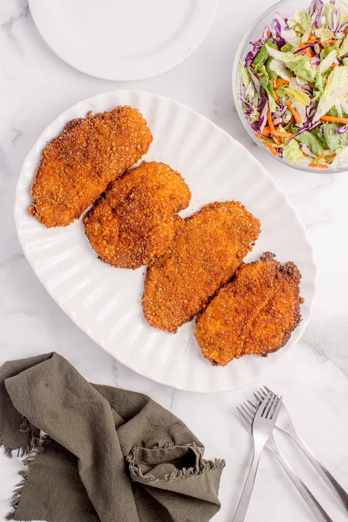 Four breaded and fried chicken cutlets are arranged on a white oval platter, with a bowl of salad, utensils, and a napkin nearby on a marble surface.