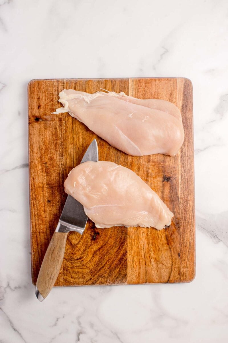 Two raw chicken breasts on a wooden cutting board, one being cut with a kitchen knife, on a white marble countertop.