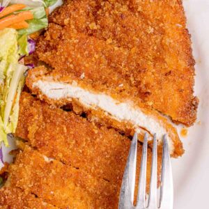 A close-up of a breaded and fried chicken cutlet partly sliced with a fork, served with a side of mixed salad on a white plate.