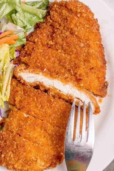 A close-up of a breaded and fried chicken cutlet partly sliced with a fork, served with a side of mixed salad on a white plate.