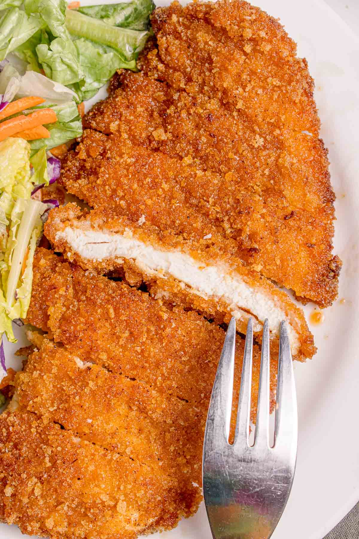 A close-up of a breaded and fried chicken cutlet partly sliced with a fork, served with a side of mixed salad on a white plate.