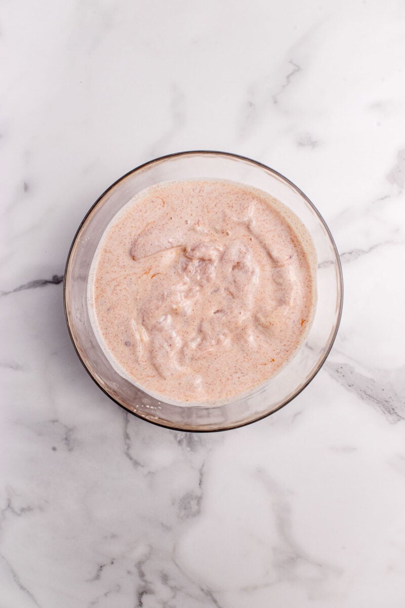 A glass bowl filled with raw chicken pieces coated in a buttermilk marinade, placed on a white marble surface.