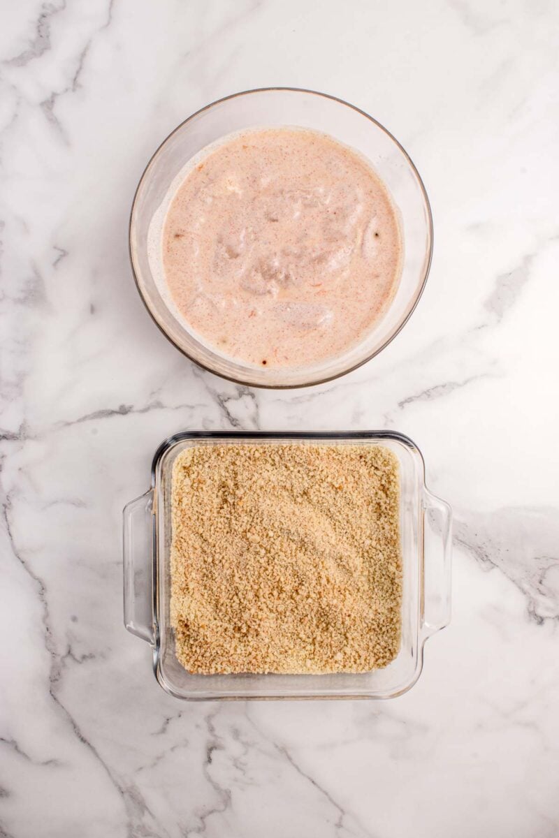A glass bowl with a seasoned marinade mixture above a square glass dish filled with breadcrumbs on a white marble surface.