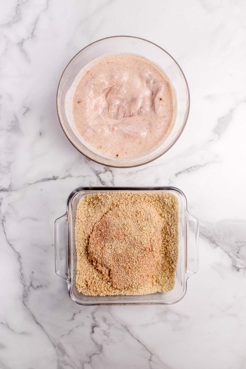 A piece of chicken is coated in a buttermilk marinade in a bowl, and another piece is being breaded in a container of breadcrumbs on a marble surface.