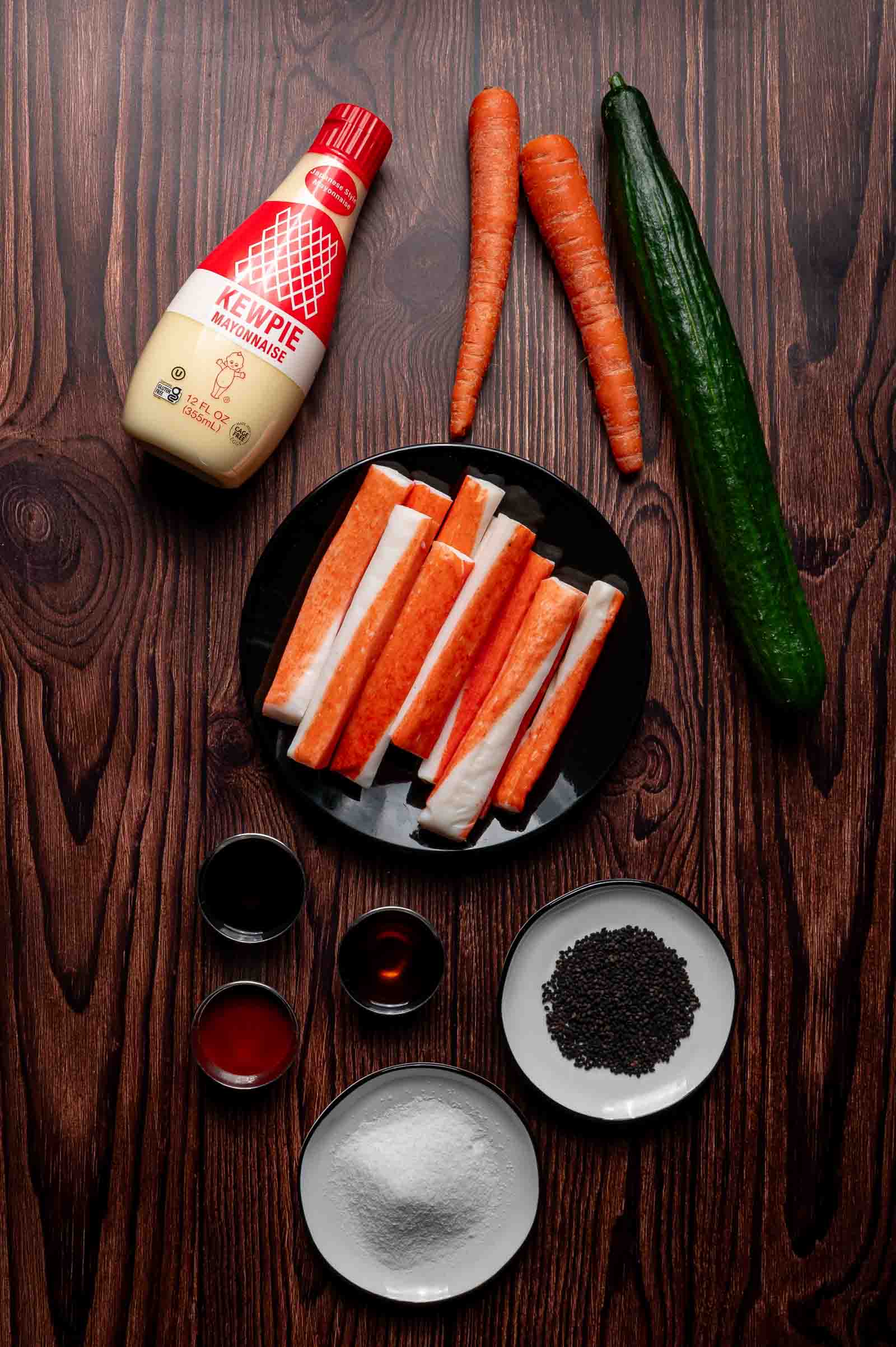 Ingredients for kani Salad on a wooden surface: Kewpie mayo bottle, two carrots, one cucumber, a black plate with imitation crab sticks, and small bowls with seasonings and spices.