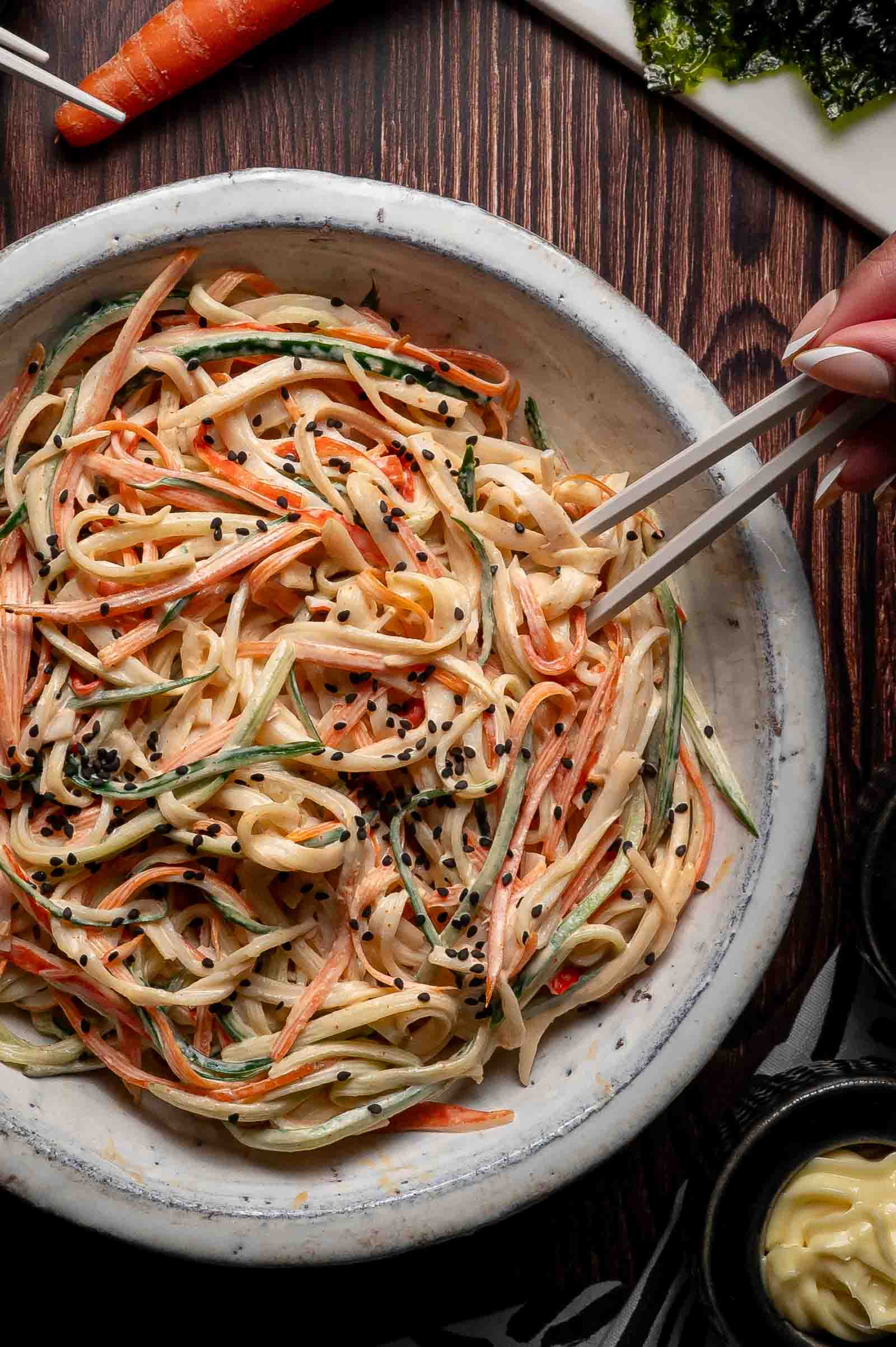 A bowl of kani salad with creamy sauce, topped with black sesame seeds. A hand uses chopsticks to pick up the noodles.