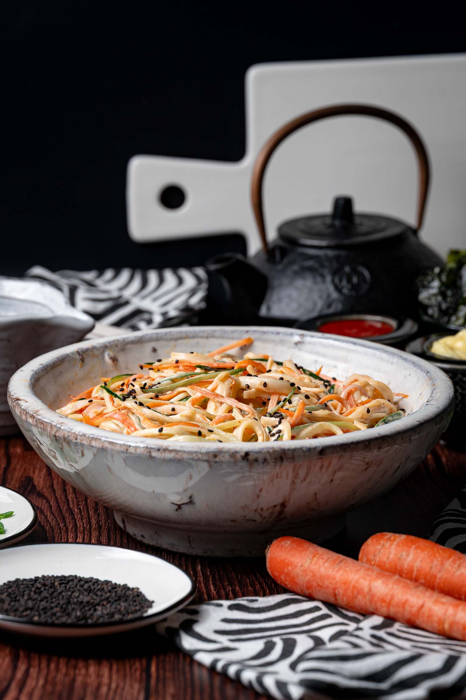 A rustic white bowl filled with kani salad topped with black sesame seeds, surrounded by carrots, a plate of seeds, and a black teapot in the background.