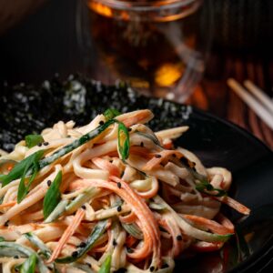 A plate of kani salad with julienned crab sticks, cucumber, green onions, and black sesame seeds, served with seaweed, with a glass of tea in the background.