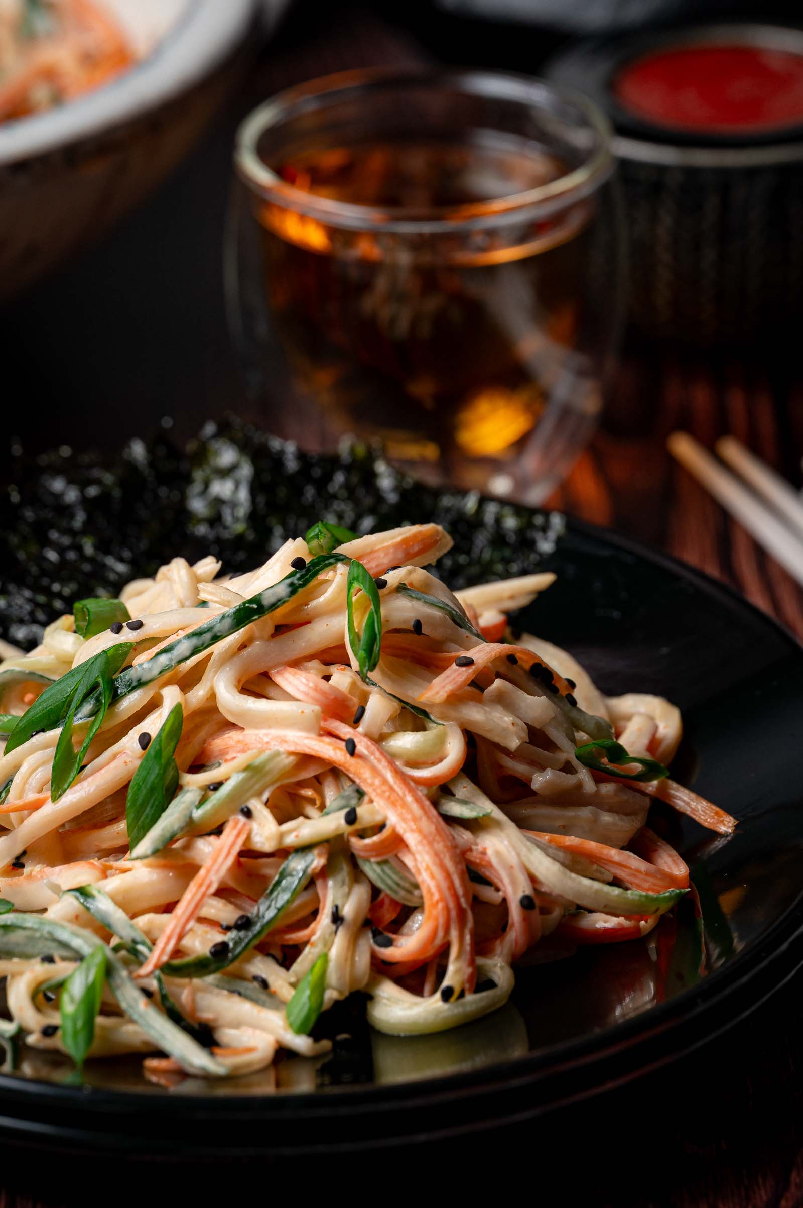 A plate of kani salad with julienned crab sticks, cucumber, green onions, and black sesame seeds, served with seaweed, with a glass of tea in the background.