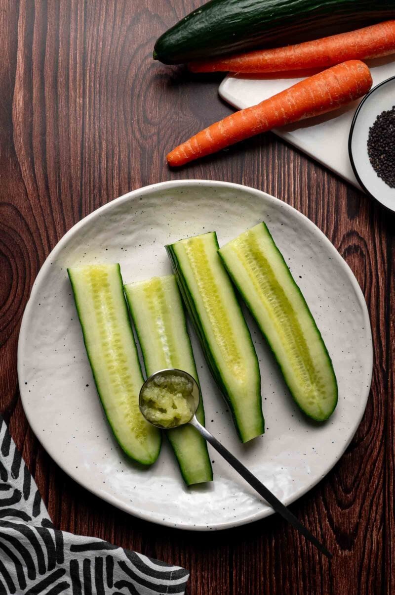 A white plate with three cucumber halves, a spoon holding cucumber pulp, a carrot, a cucumber, and a bowl of seeds on a wooden table.