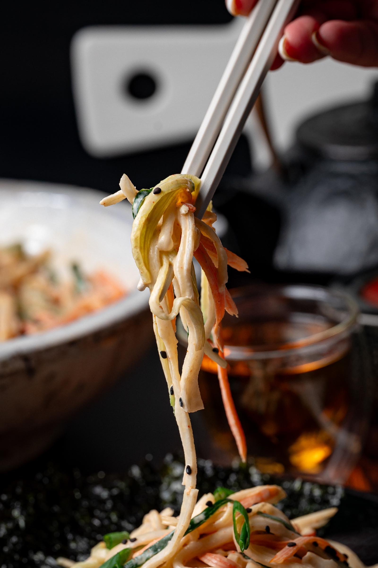 Chopsticks holding up a portion of kani salad with vegetables and black sesame seeds, with a bowl and drink in the background.