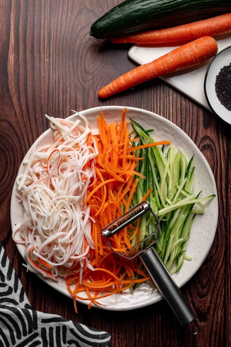 Oval plate with shredded surimi, julienned carrots, and julienned cucumbers on a wooden table, with whole carrots, a cucumber, and a julienne peeler nearby.