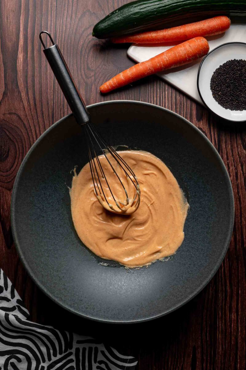 A black bowl with a whisk mixing a creamy light orange dressing, placed on a wooden surface beside carrots, cucumber, and a plate of black sesame seeds.