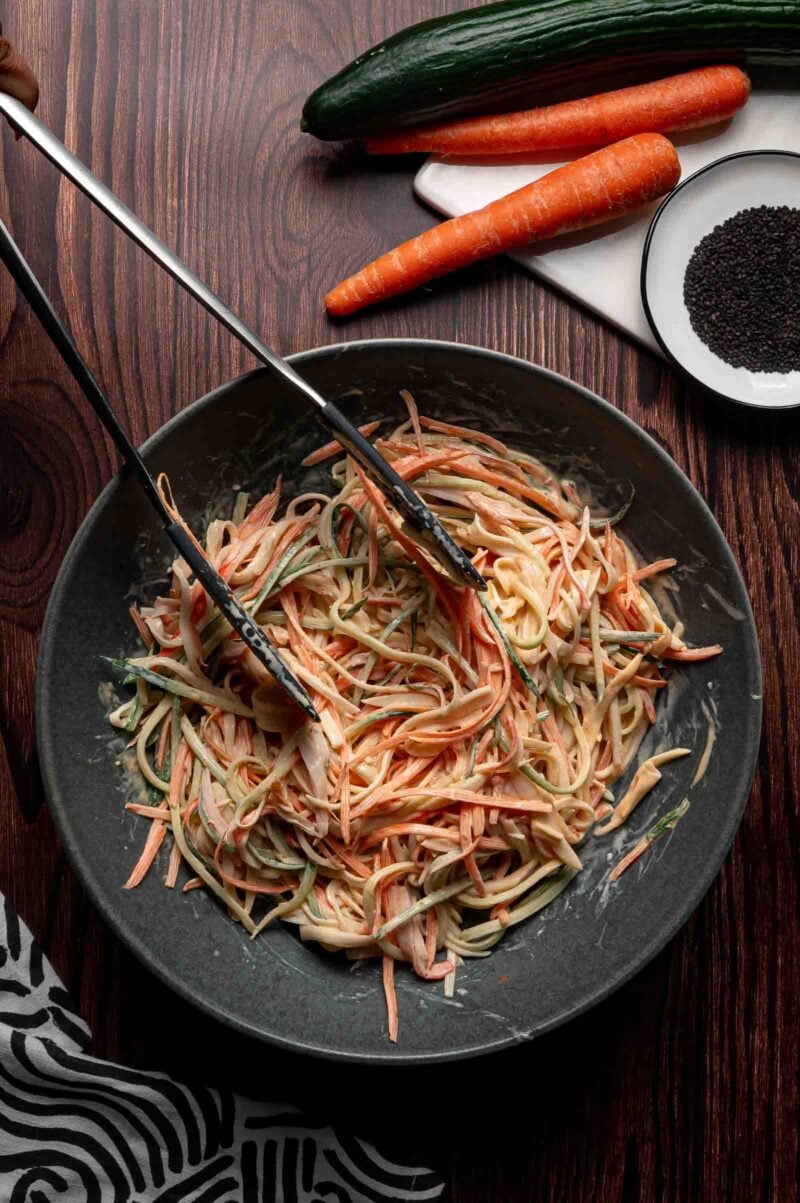 A bowl of creamy kani salad with crab, shredded carrots and cucumber being tossed with tongs; whole carrots, cucumber, and a plate of black sesame seeds are nearby.
