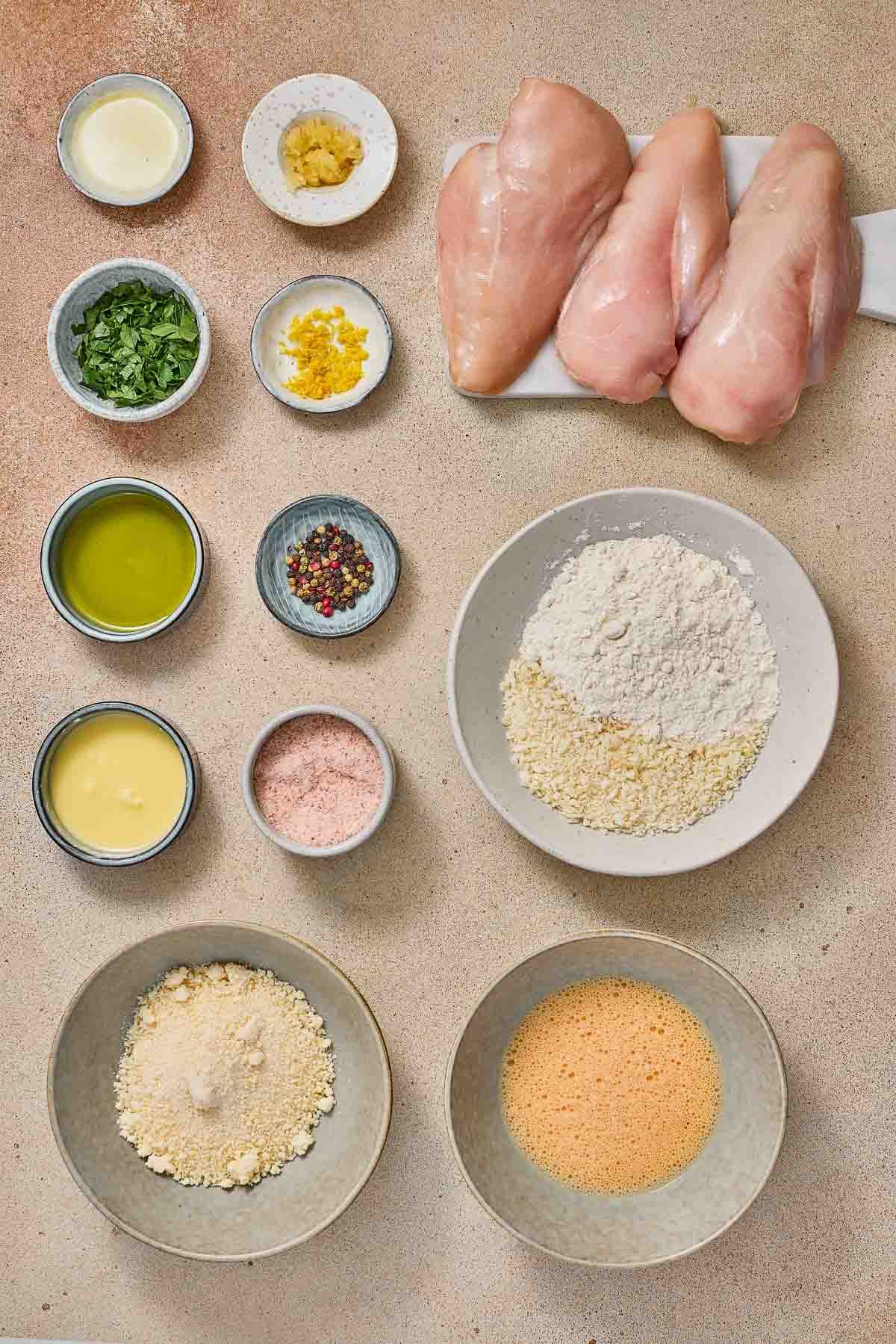 Raw chicken breasts, herbs, spices, flour, breadcrumbs, eggs, oil, and seasoning arranged in small bowls on a countertop, prepared for a recipe.