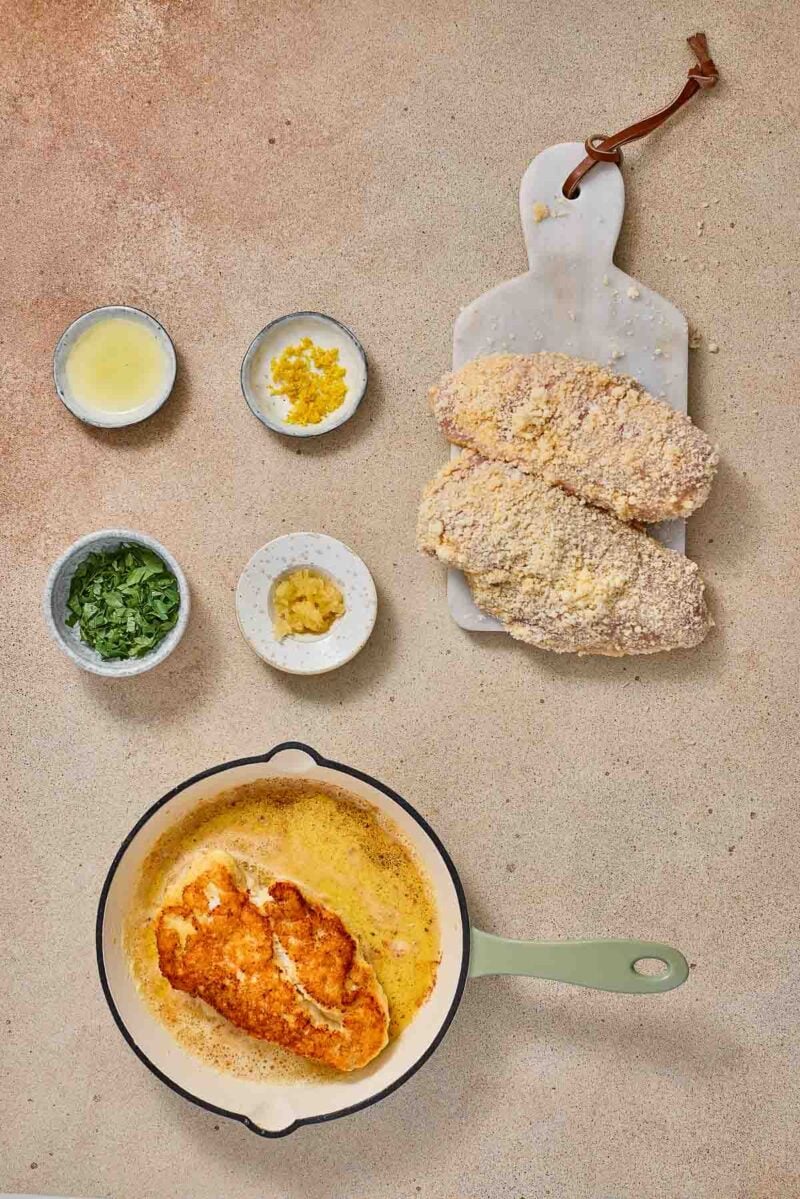 A pan with breaded chicken cooking in butter and oil, next to a marble board with raw breaded chicken and small bowls of lemon juice, zest, garlic, and chopped parsley.