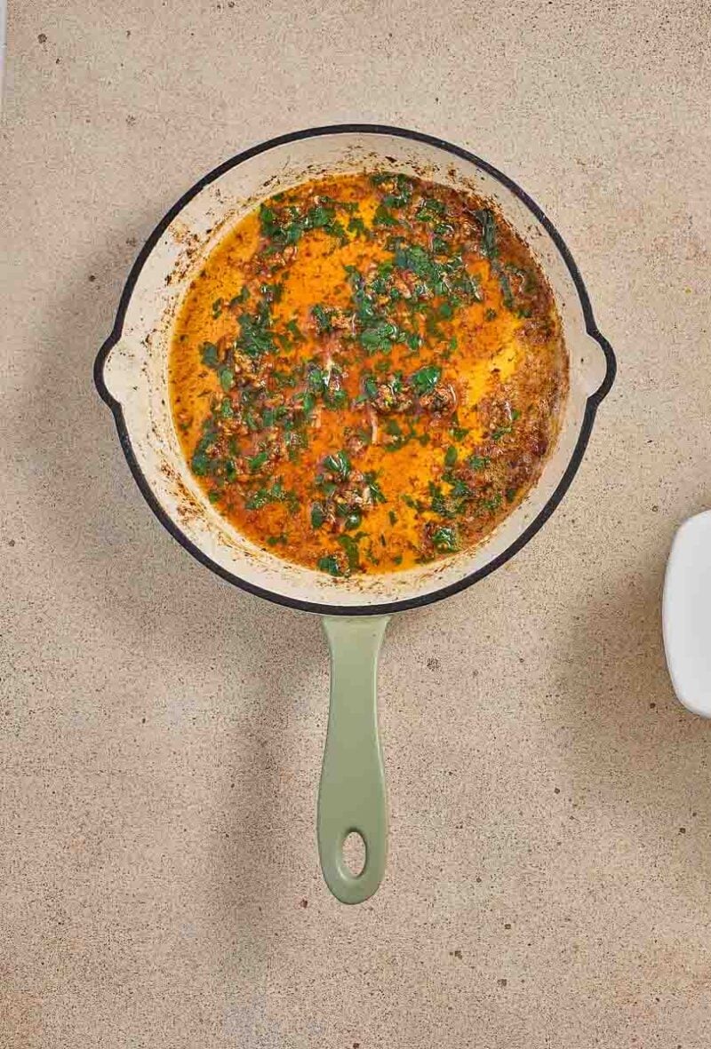 Overhead view of three cooked chicken breasts in a white baking dish, a pan of buttery lemon sauce, and a small bowl of chopped herbs on a beige surface.