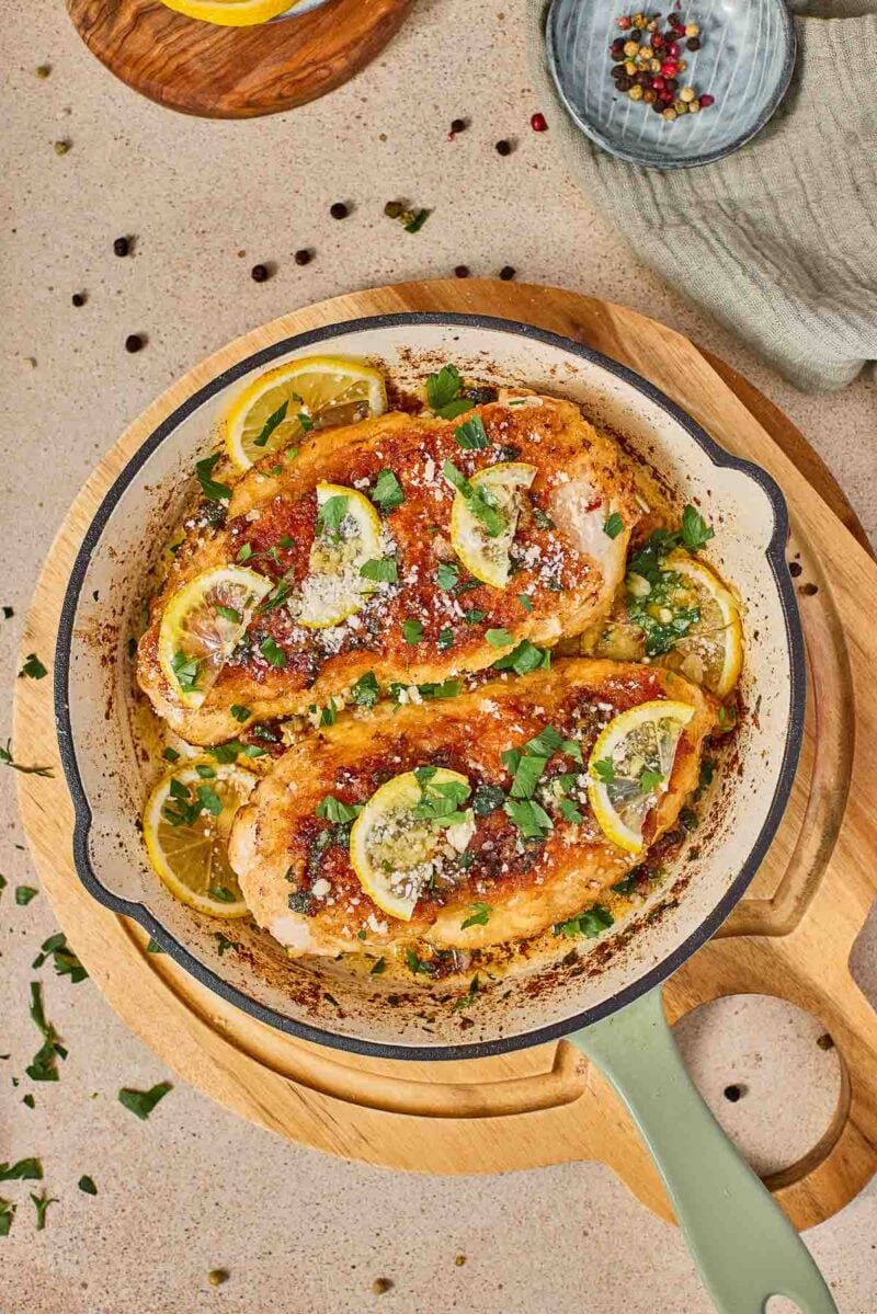 A skillet with cooked chicken romano topped with lemon slices, herbs, and grated cheese, placed on a wooden board.