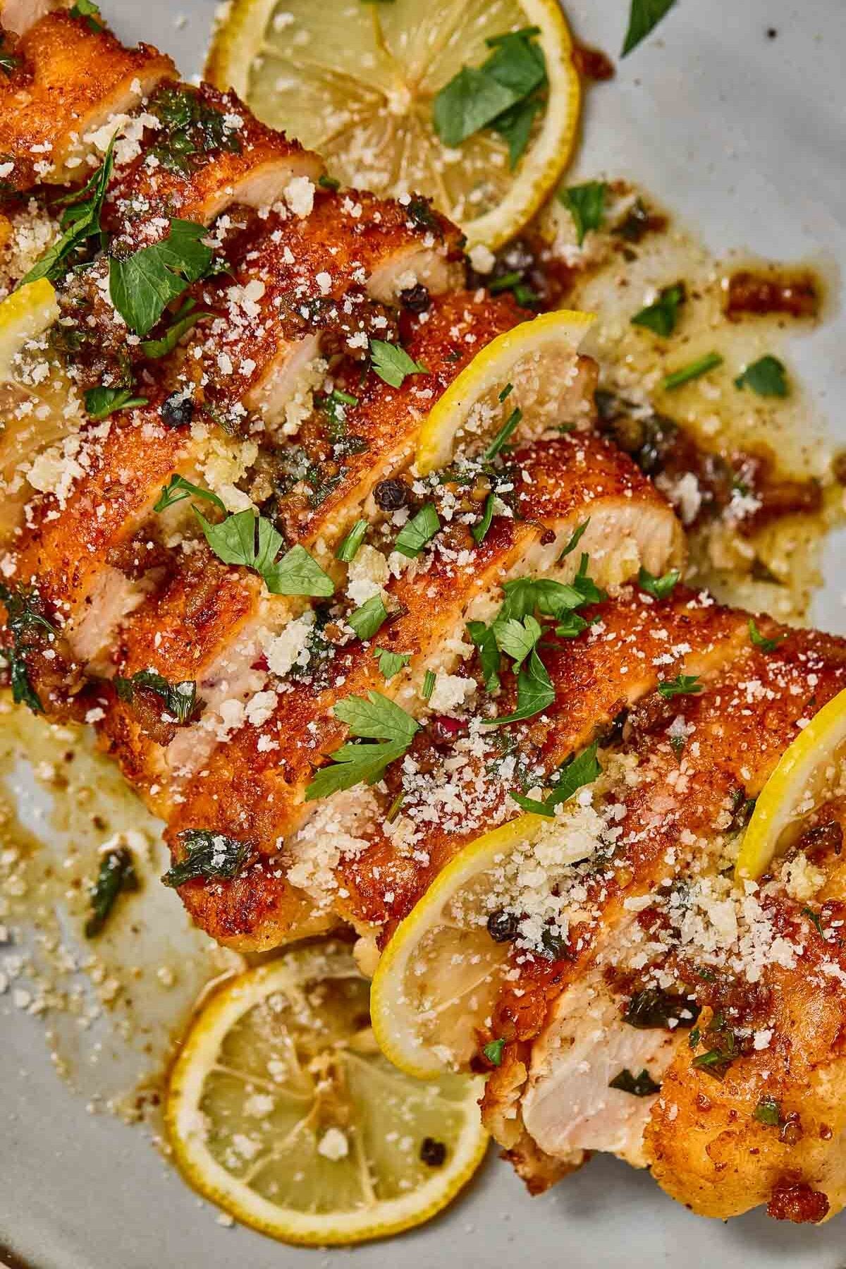Sliced cooked chicken romano garnished with lemon slices, chopped parsley, grated cheese, and seasoning on a plate.