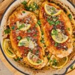 Chicken romano in a skillet, topped with lemon slices, grated cheese, and chopped parsley, with lemon slices lining the pan.