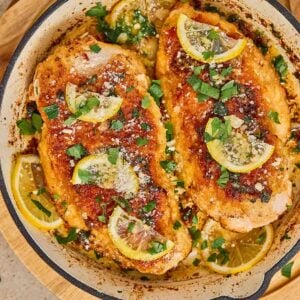 Chicken romano in a skillet, topped with lemon slices, grated cheese, and chopped parsley, with lemon slices lining the pan.
