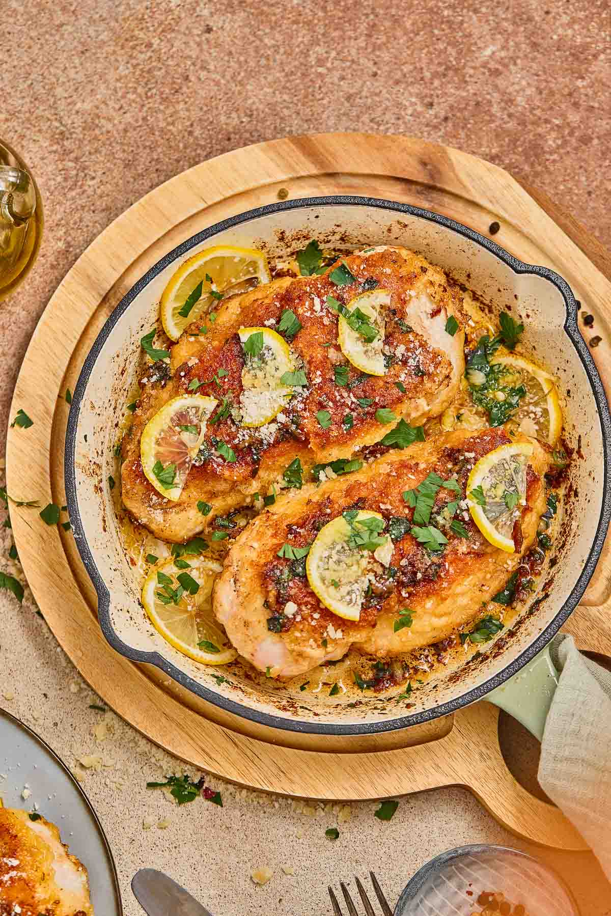 Two cooked chicken breasts in a skillet, garnished with lemon slices and chopped herbs, served on a wooden trivet.