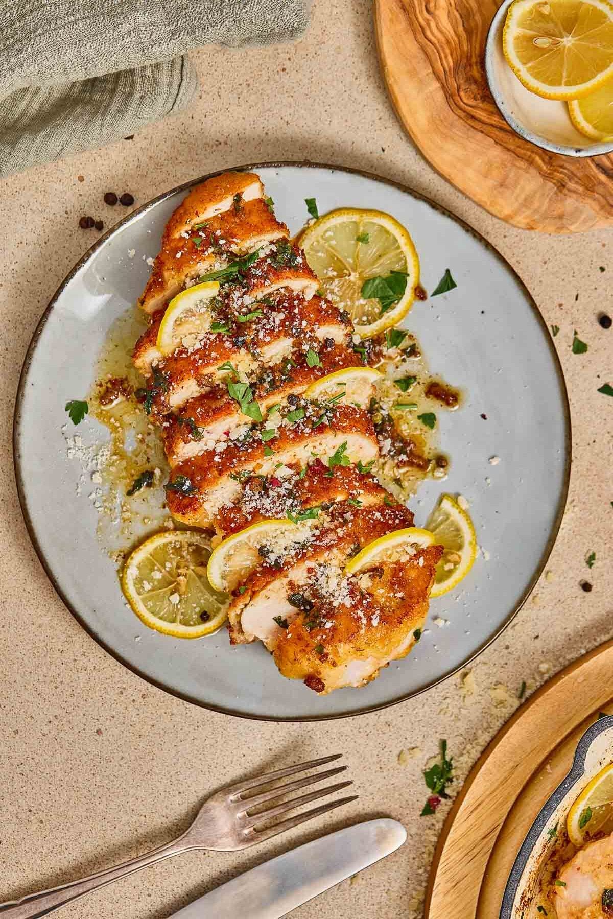 Sliced cooked chicken romano garnished with lemon slices, herbs, and grated cheese on a gray plate, with a fork and additional lemon slices nearby.