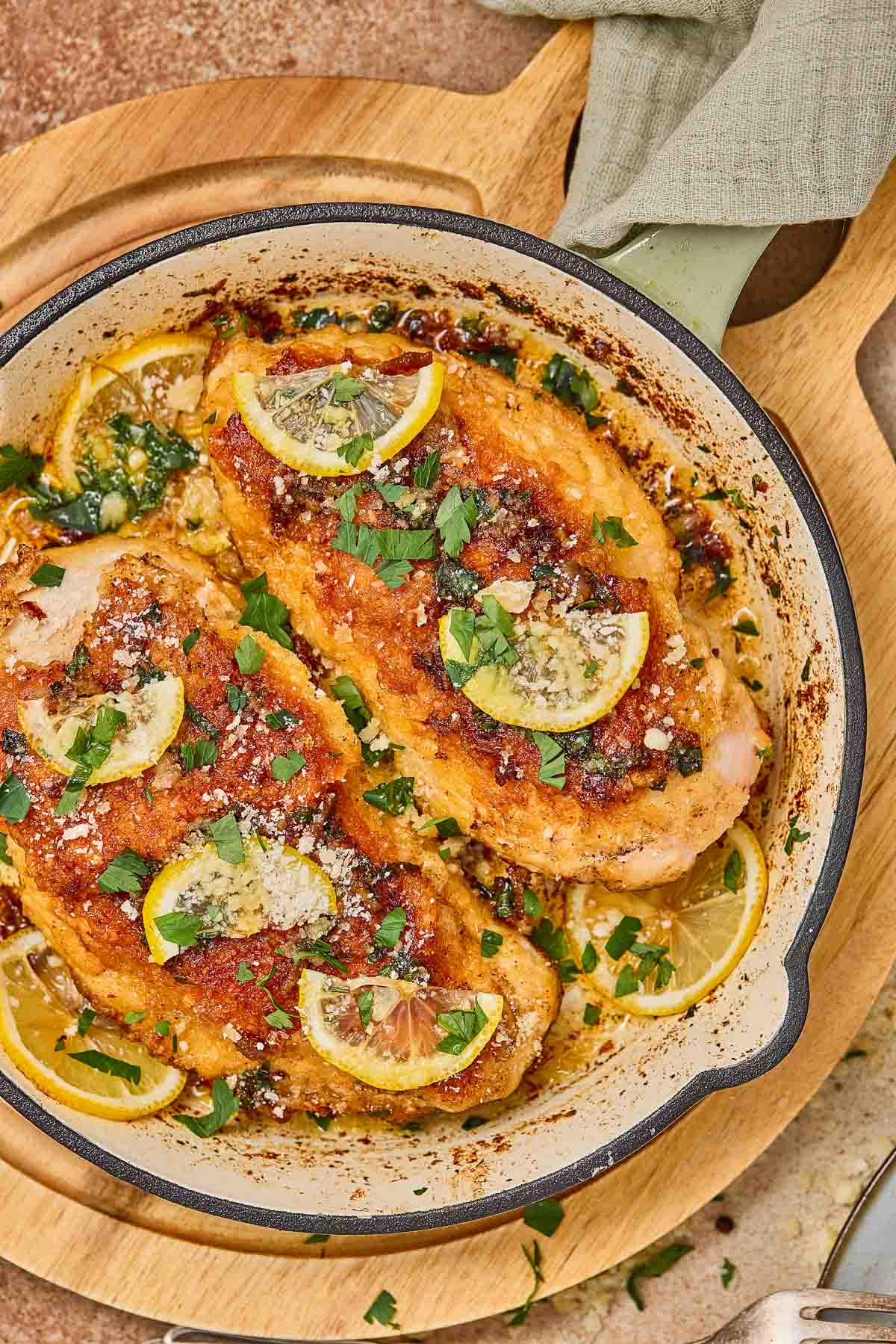 Two cooked romano chicken breasts in a skillet, topped with lemon slices, fresh parsley, and grated cheese, on a wooden board.