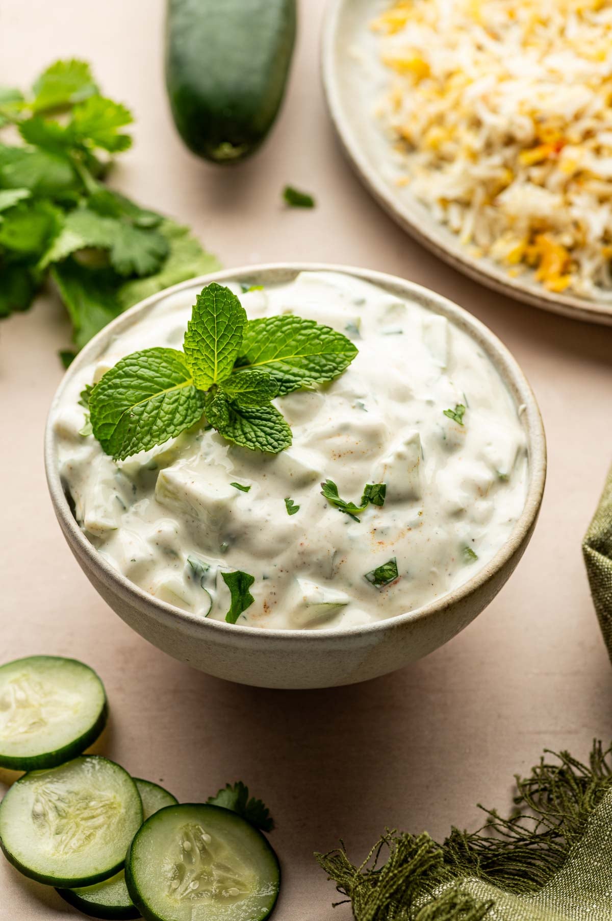 A bowl of cucumber raita garnished with mint leaves, with sliced cucumbers, cilantro, and a plate of rice in the background.