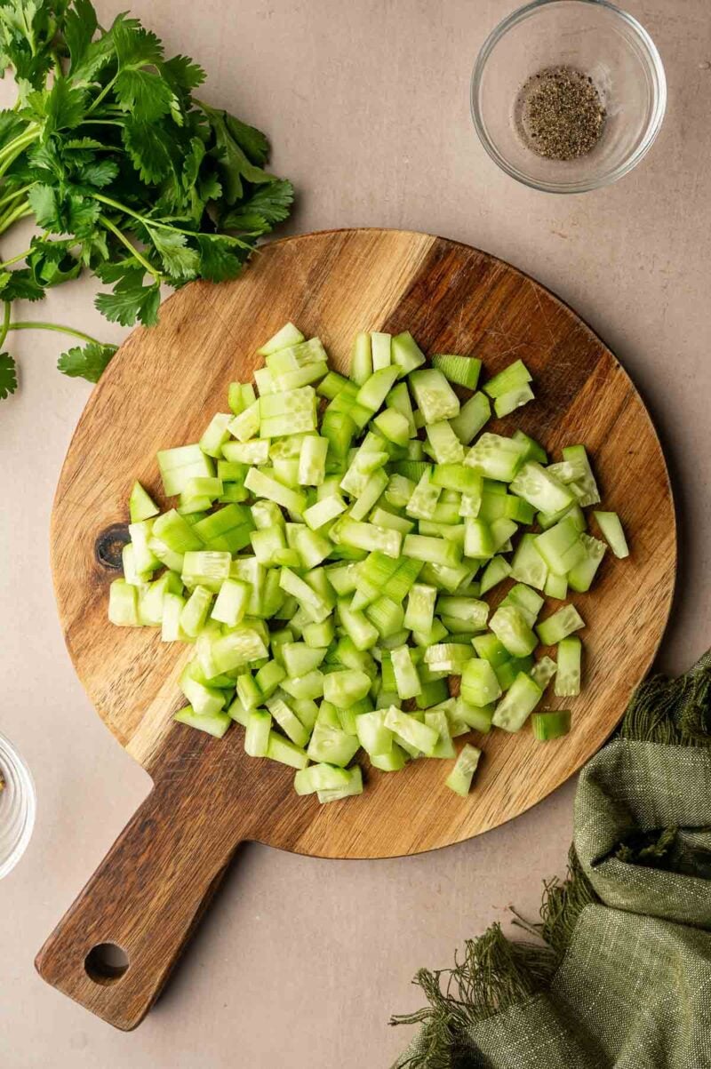 Diced cucumber on a wooden cutting board, with fresh cilantro, a bowl of ground pepper, and a green cloth nearby.