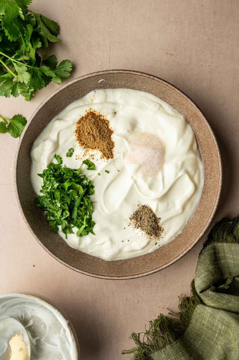 A bowl of yogurt topped with chopped cilantro, ground cumin, black pepper, and salt, with fresh cilantro and a green cloth nearby.