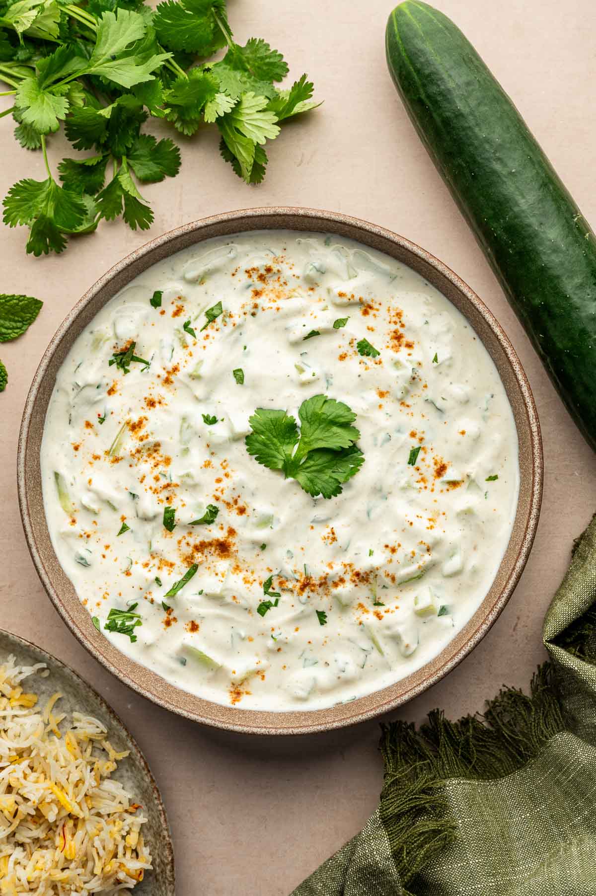 A bowl of cucumber raita garnished with cilantro and spices, surrounded by fresh cilantro, a cucumber, and a plate of rice.