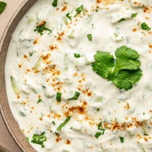 A bowl of creamy raita garnished with chopped herbs, ground spices, and a sprig of cilantro, with a partial view of rice and herbs in the background.
