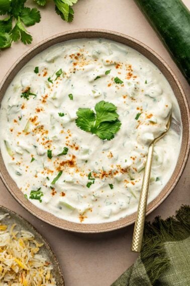 A bowl of cucumber raita garnished with cilantro leaf, chopped herbs, and a sprinkle of spices, with a gold spoon resting inside. Rice, cilantro, and cucumber are nearby.