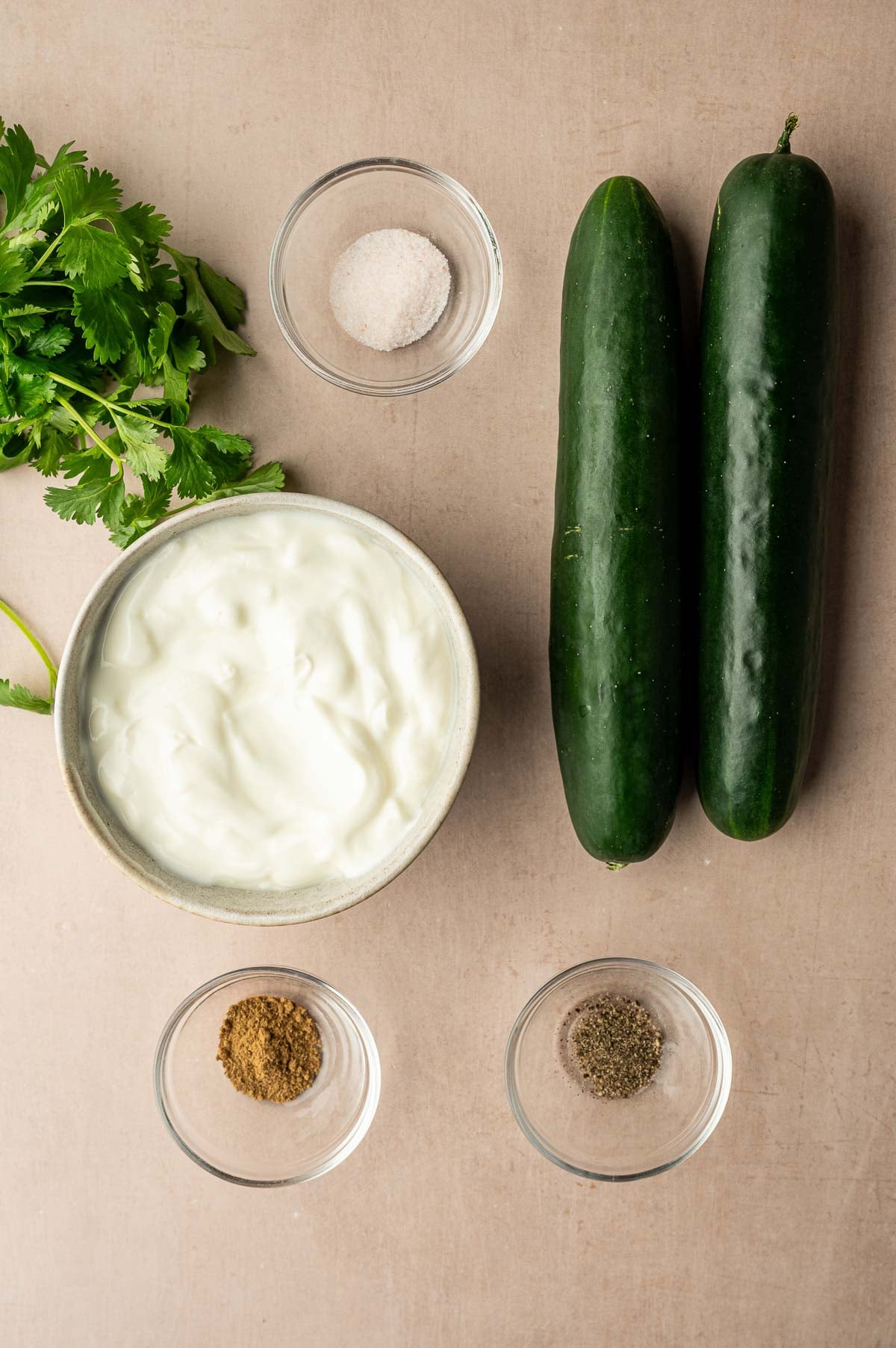 A bowl of yogurt, two whole cucumbers, fresh cilantro, and small glass bowls containing salt, ground cumin, and ground black pepper are arranged on a neutral surface.