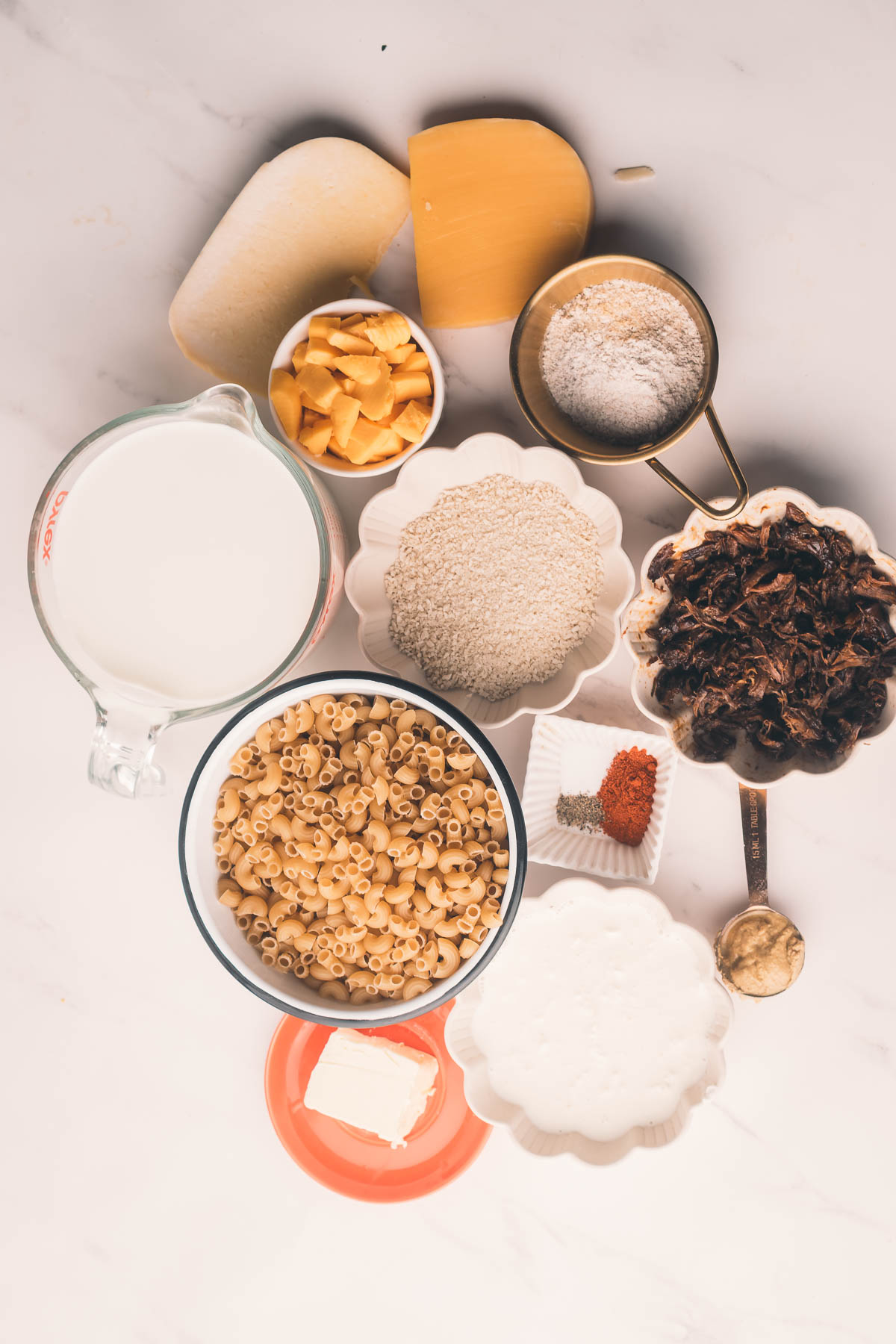Various ingredients for a mac and cheese dish are arranged on a white surface, including uncooked macaroni, cheese, milk, flour, cooked mushrooms, spices, butter, and cream.