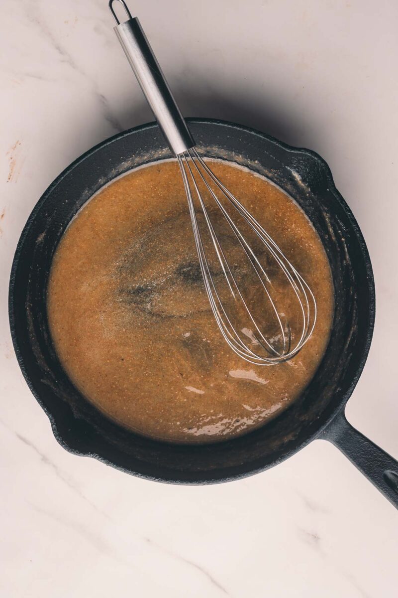 A whisk rests in a cast iron skillet containing brown roux on a white marble surface.
