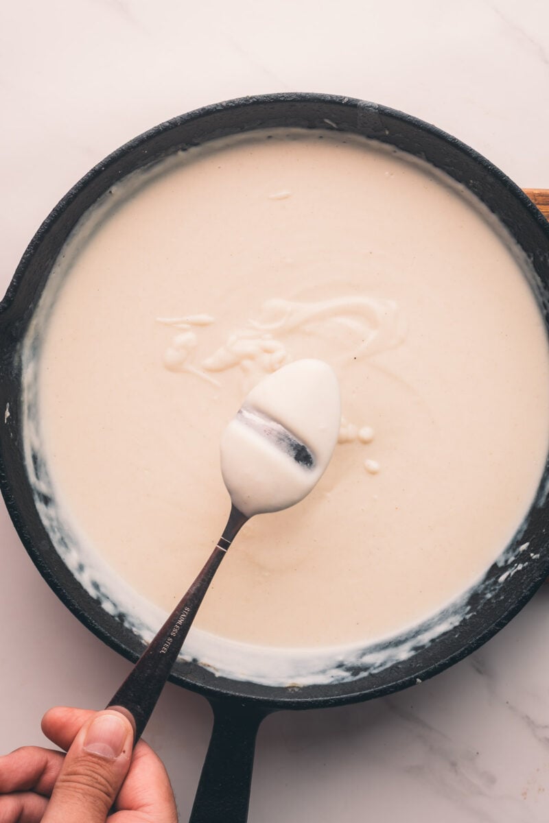 A hand holds a spoon coated with creamy white sauce above a cast iron pan filled with the same white sauce on a light surface.