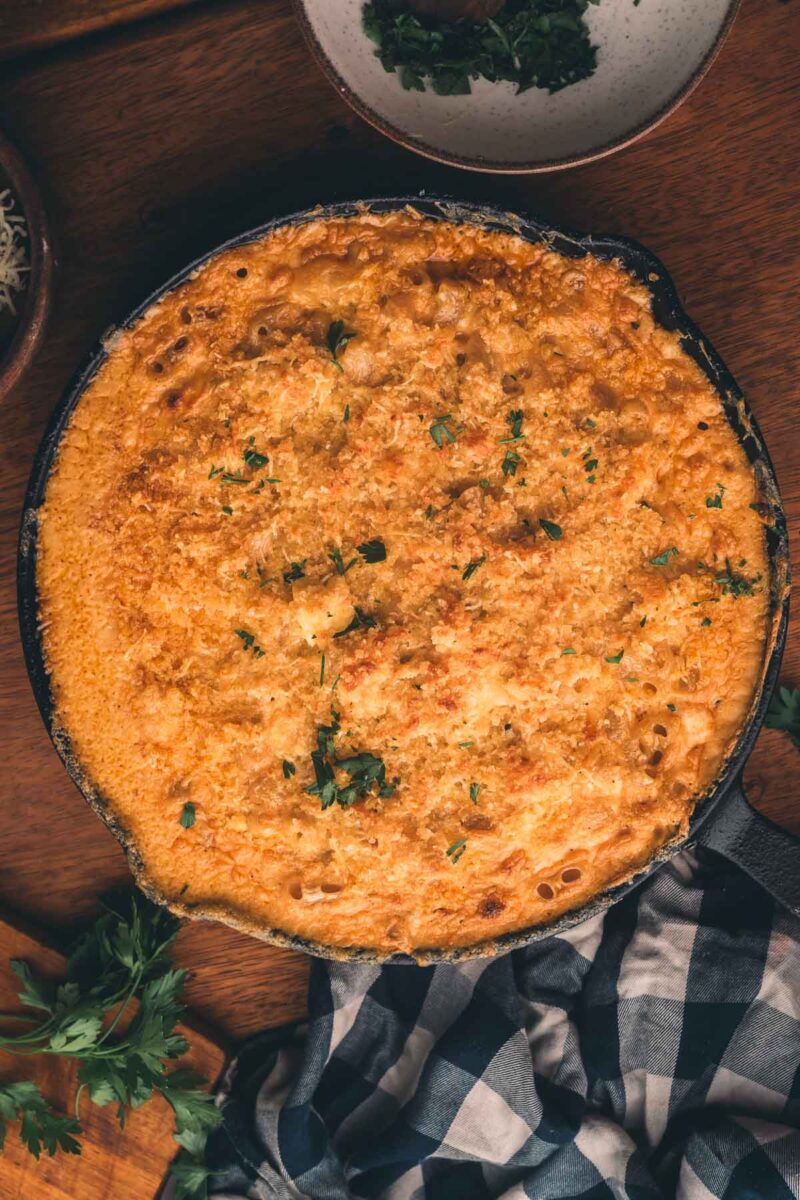 A baked macaroni and cheese dish in a cast iron skillet, topped with a golden brown crust and garnished with chopped parsley.