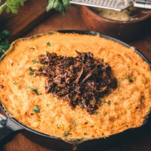A cast iron skillet filled with baked macaroni and cheese, topped with shredded braised beef and garnished with chopped parsley.
