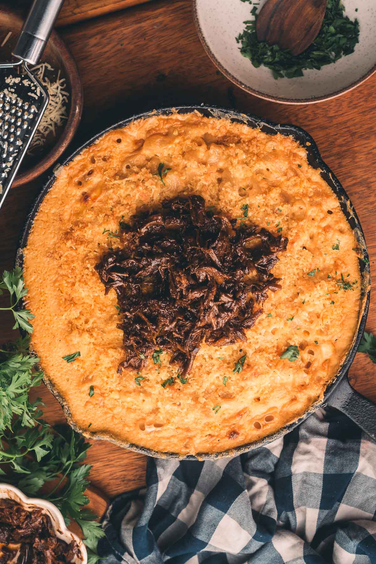 A cast iron skillet filled with baked macaroni and cheese, topped with shredded beef, sits on a wooden table with herbs and a cheese grater nearby.