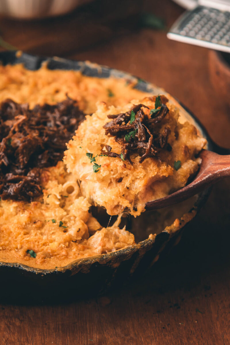 A wooden spoon scoops baked macaroni and cheese topped with leftover braised beef from a cast iron skillet on a wooden table.