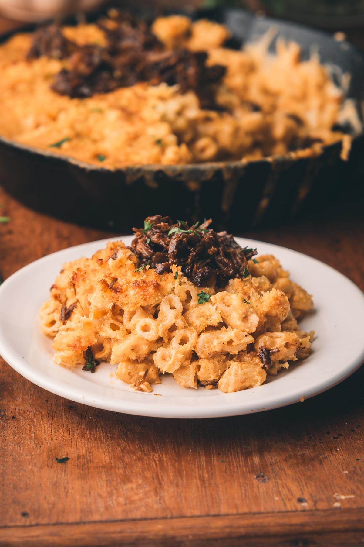 A serving of baked macaroni and cheese topped with braised beef shanks on a white plate, with a skillet of macaroni and cheese in the background.